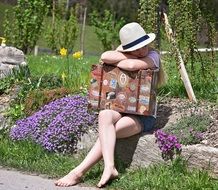 Girl sitting with suitcase in the garden with colorful flowers