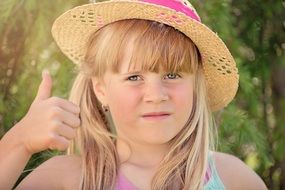 Portrait of a blonde girl in pink hat with thumb up in light