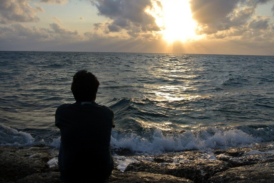 man sits on stones near the ocean at sunset