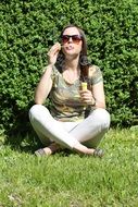 A girl sits under a green bush and blows soap bubbles