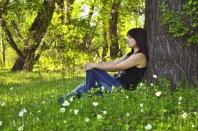 Girl sitting under the green tree on the beautiful green grass with white flowers