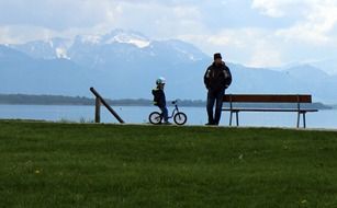 dad at the bench and the child on the bike on the lake