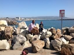 People sit on boulders on the seashore with mountains