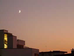 new moon in evening sky above roofs