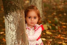 baby girl in a dress looking from behind a tree