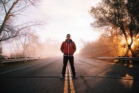 young man in a red jacket stands on the dividing strip on the highway