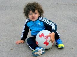 A child with curly hair with a ball sits on the asphalt