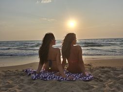 two young long haired girls sitting on sand at sea