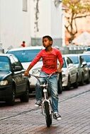 dark skin child boy riding bicycle on street along cars in row