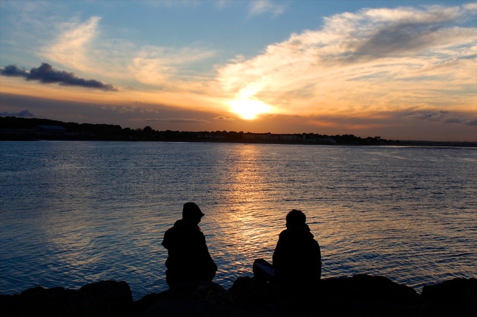 two persons sit on stones in front of water at sunset