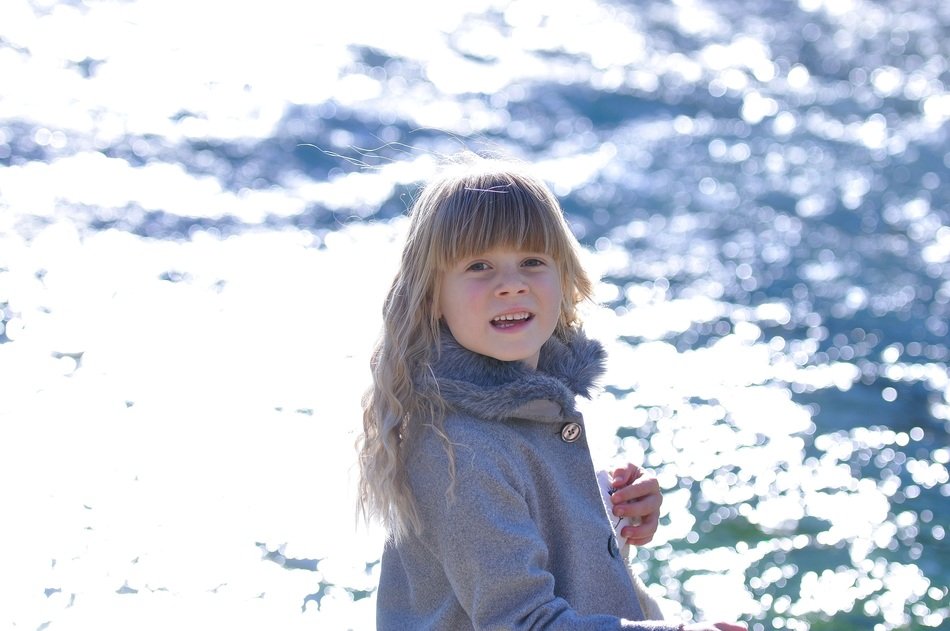 blond child girl in blue coat in front of glossy water
