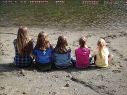 back view of five child girls sitting in row on sand at water