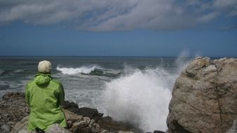 person in rain jacket sitting on rock at stormy sea, south africa