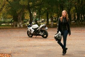 blonde girl in a leather jacket with a helmet in hand on the background of a motorcycle