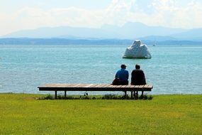 two persons sitting on wooden bench at calm water