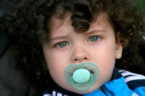 the curly baby with blue eyes and pacifier in his mouth