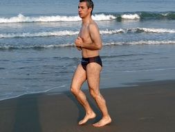 a man in swimming trunks jogging along the shore