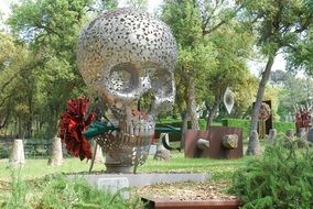 silver skull with red carnation in teeth, modern metal sculpture in park, spain