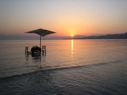 table and chairs beneath parasol in water at sunrise, greece, corfu, roda