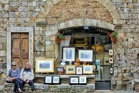 happy old woman and man sitting at old stone wall in front of entrance to photo shop, italy, sant gimignano