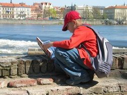 student with a backpack reading a book on the embankment in Prague