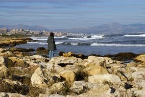 woman stays on rocky coast in view of city at sea, Spain, Alicante