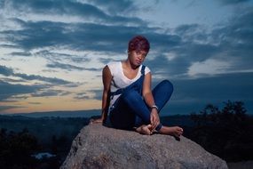 girl in denim overalls sits on a stone at dusk