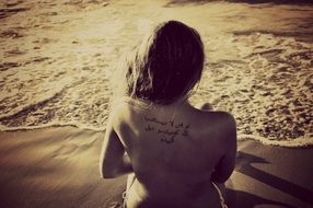 Young Woman with tattoo on back sits on sand at water