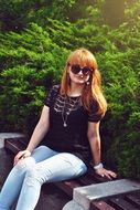 girl in glasses sits near the green bush