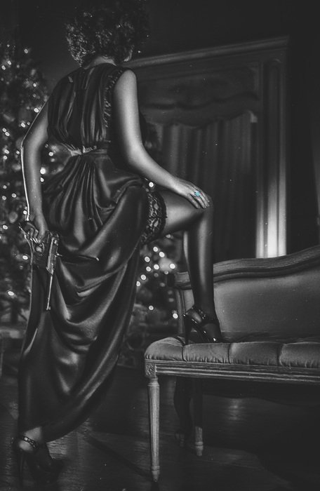 black and white picture of a girl in a black dress with the foot on the chair near the mirror