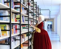 Woman in white wig and red vintage dress stands at shelves with books in library