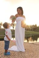 child boy gives bouquet to happy Pregnant Woman in long white dress