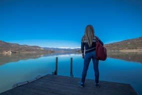 young blonde Girl looking at beautiful mountain landscape from pier