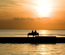 couple is sitting on a bench on a pier against the sunset