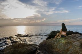 young girl sits on stone in front of sea at Sunset