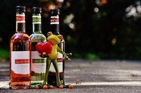 toy frog holds heart in front of three bottles of wine