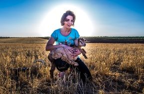 young woman sits on harvested field with goat in hands
