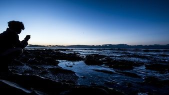 Person taking photo of sea at dusk with smartphone, silhouette