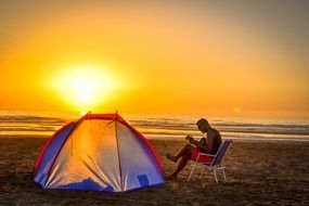 man playing Guitar on beach beside of tent at colorful sunset on horizon