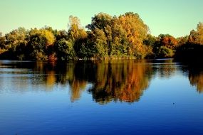 trees are reflected in the blue water of the lake in autumn