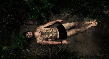 young man in underpants lays on his back in mud