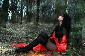 girl in black clothes and with a red cape sits on the ground in the forest