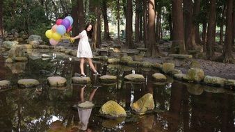 nice smiling asian child girl with colorful balloons walking by stones through pond in park