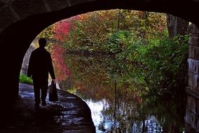 Silhouette of a person on a trail under a bridge over a water channel with beautiful and colorful plants