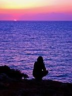 silhouette of a man squatting on the shore of the blue sea on a background of pink sunset