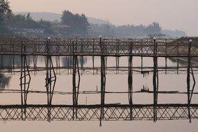 old bamboo bridge mirroring on calm water in foggy landscape, india