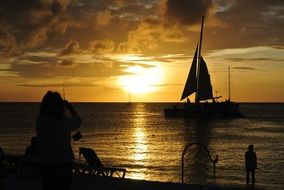 Silhouettes of people watching sailboat on the water at colorful sunset background