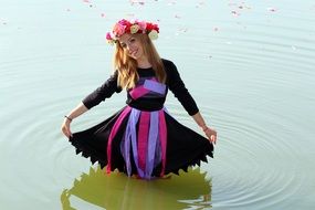 A girl in a dark dress and a wreath on her head stands in the water