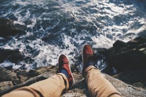 legs of a young man sitting on a cliff on the background of sea foam