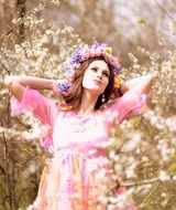 young woman with colorful flower wreath in pink dress among the flowers